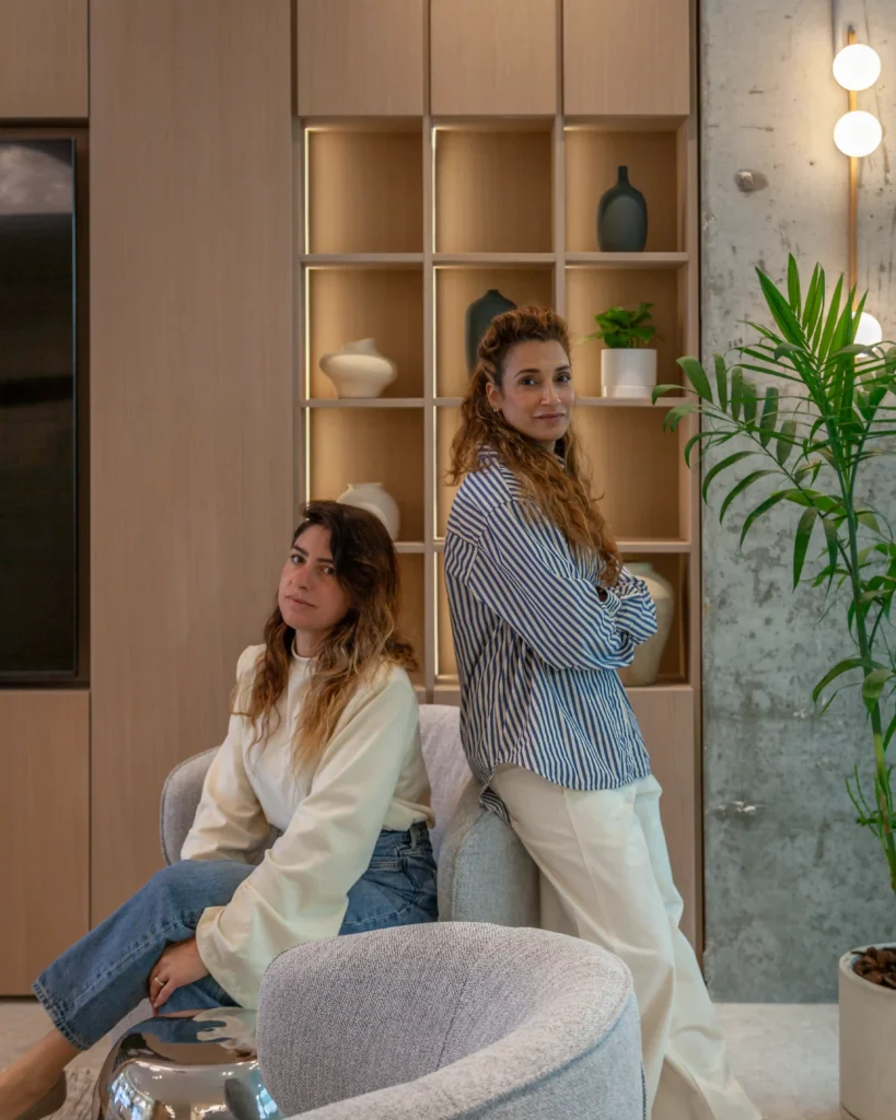 מעצבות הפנים דנה וענת מסטודיו DADA. תמונת פורטרט מתוך פרויקט עיצוב דופלקס בהרצליה המאופיין במינימליזם חם, רקע ספריית עץ אלון ונגיעות ירוקות.